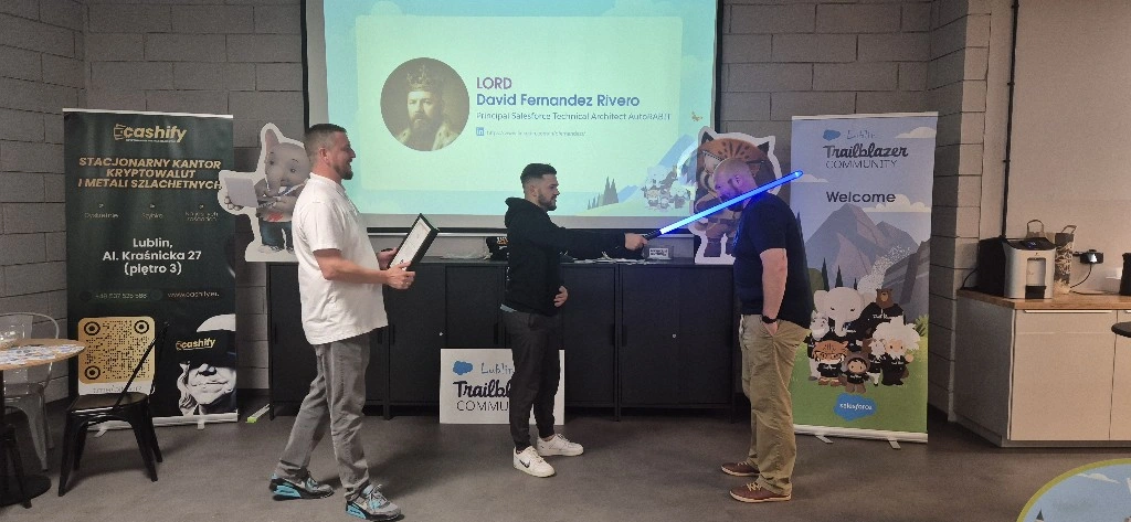 Trzech mężczyzn podczas prezentacji, jeden z nich trzyma miecz świetlny, a na ekranie wyświetlana jest prezentacja.