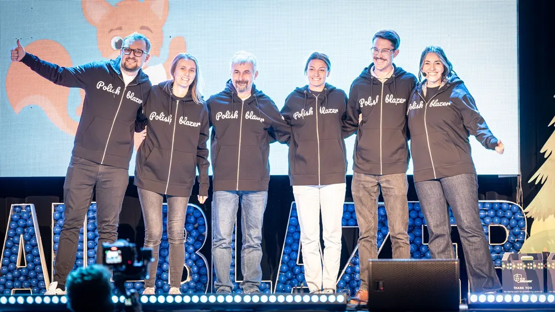 Grupa osób na scenie stoi ramię w ramię w czarnych bluzach z białym napisem Polish, uśmiechnięci, za nimi duża, kolorowa iluminacja i cyfrowa dekoracja